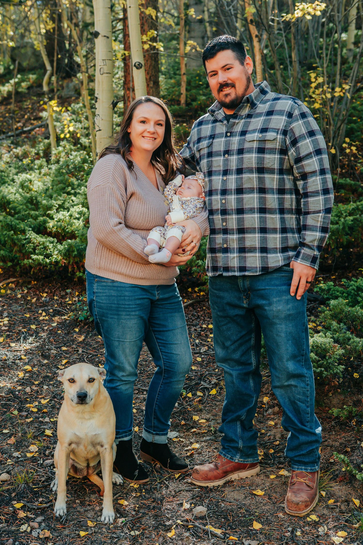 A couple poses with their baby and dog in an autumn forest setting.