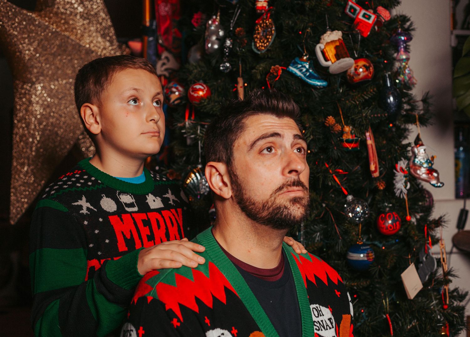 Two people wearing festive Christmas sweaters pose in front of a decorated tree.