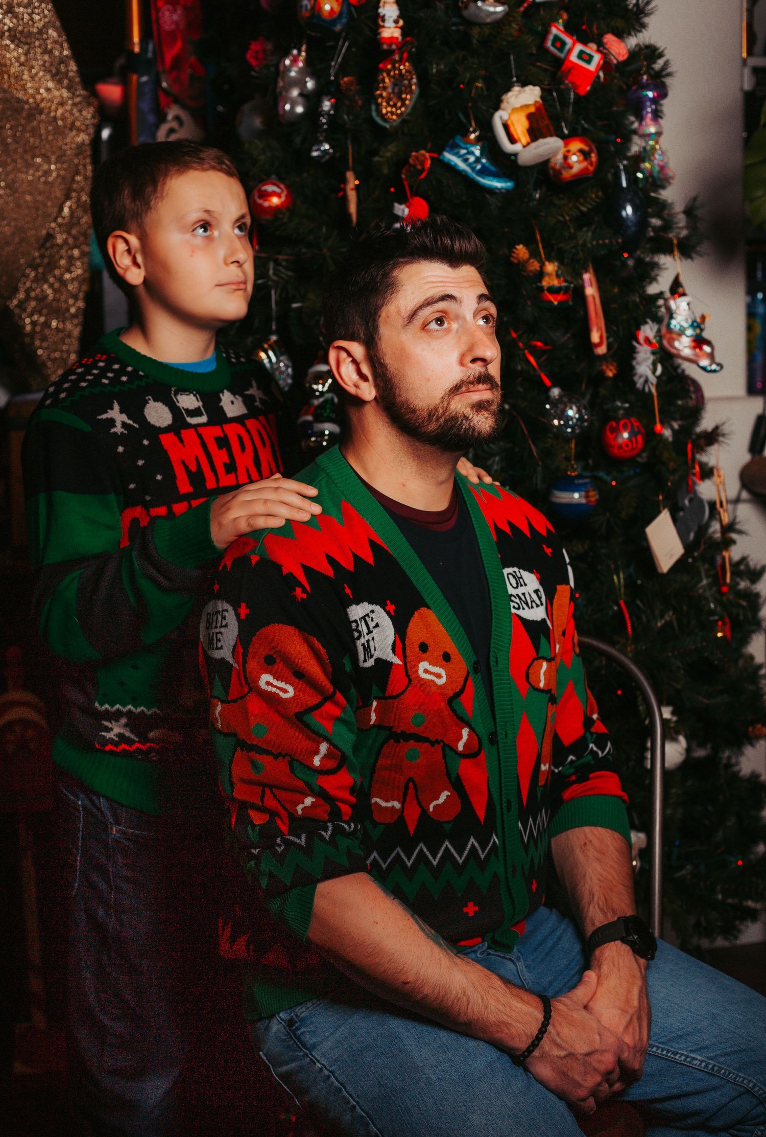 Two people in ugly Christmas sweaters pose seriously in front of a decorated tree.