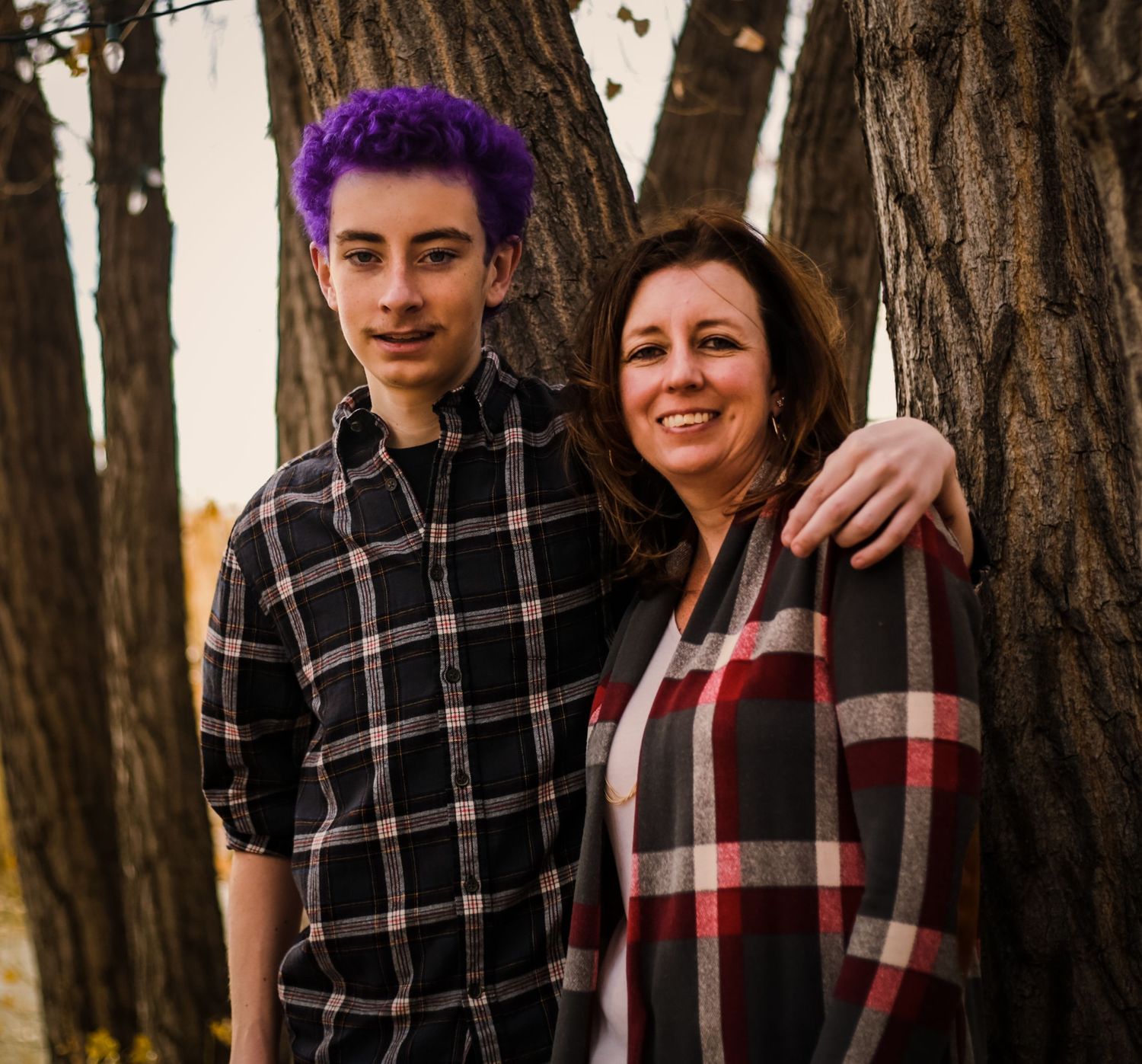 Mother and son with purple hair wearing plaid shirts pose between trees.