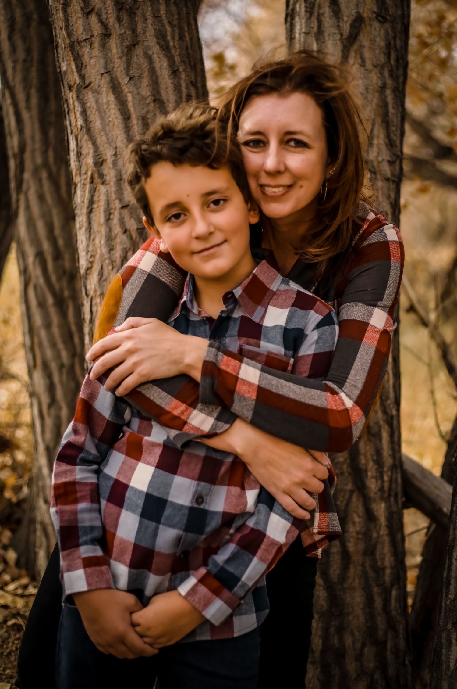 Mother and son wearing matching plaid shirts embrace while standing between trees.