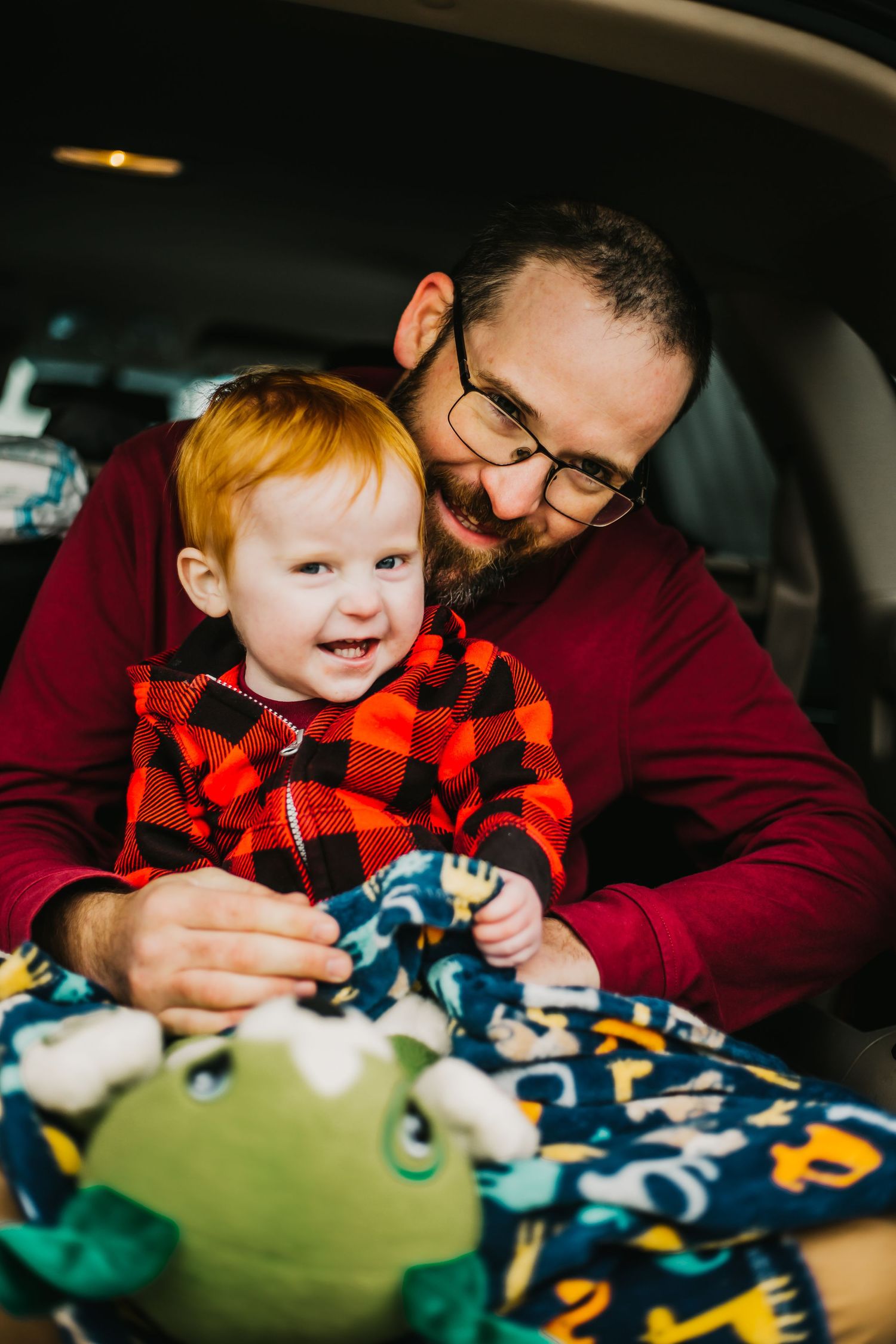 A parent in red sweater plays with toys while sitting with a toddler wearing a buffalo plaid shirt.