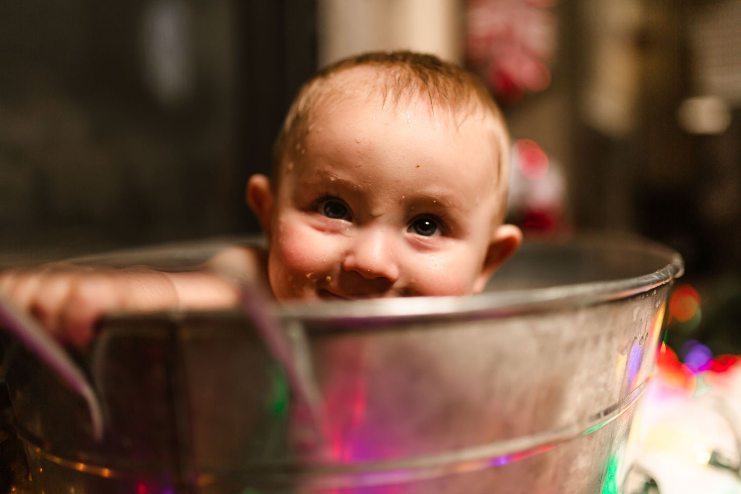 Happy baby sitting in a shiny metal mixing bowl with Christmas lights in background.