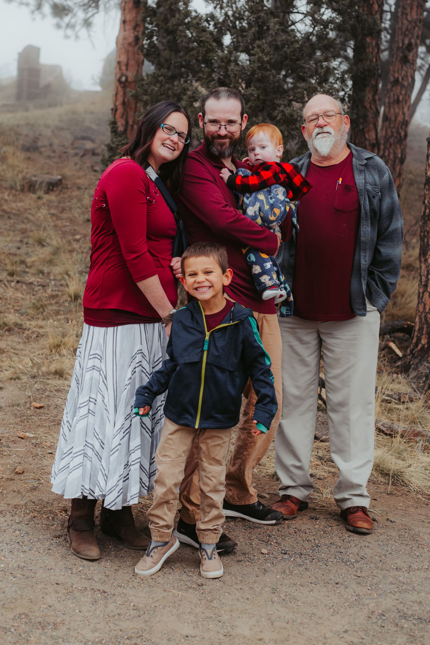A family dressed in coordinating red outfits poses together on a misty hiking trail among pine trees.