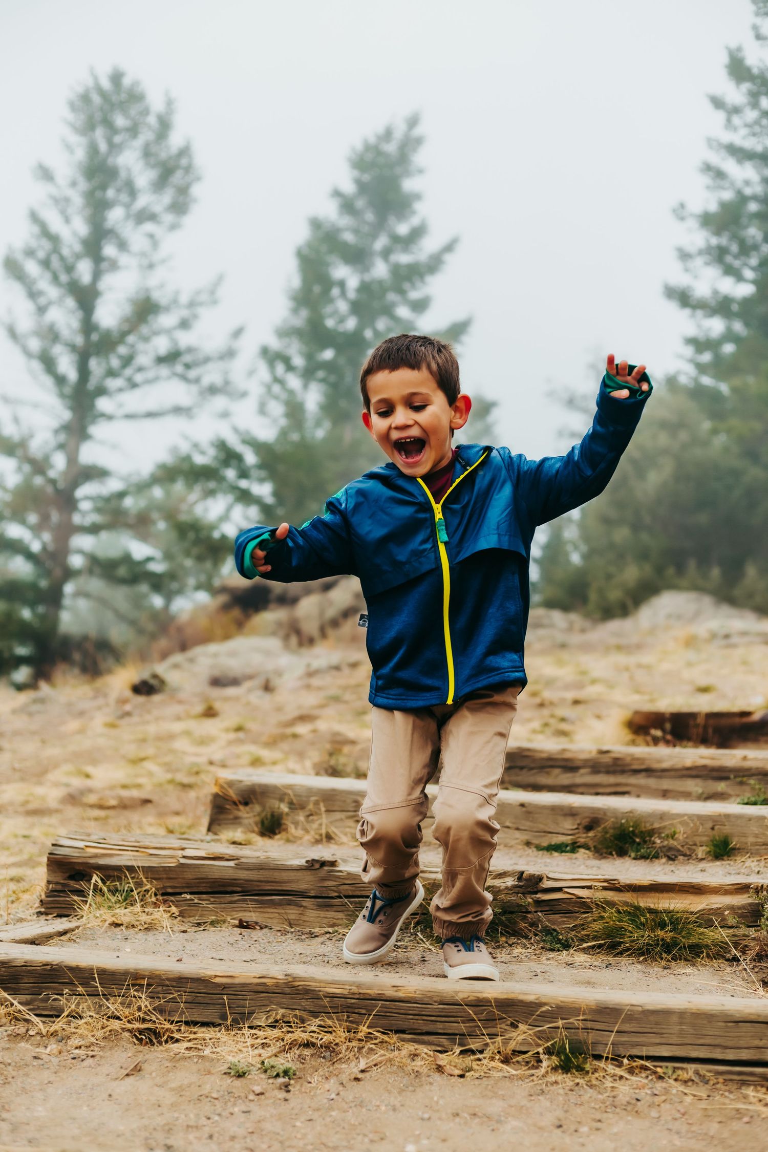 Young child in blue jacket jumping joyfully on wooden steps in a misty forest setting.