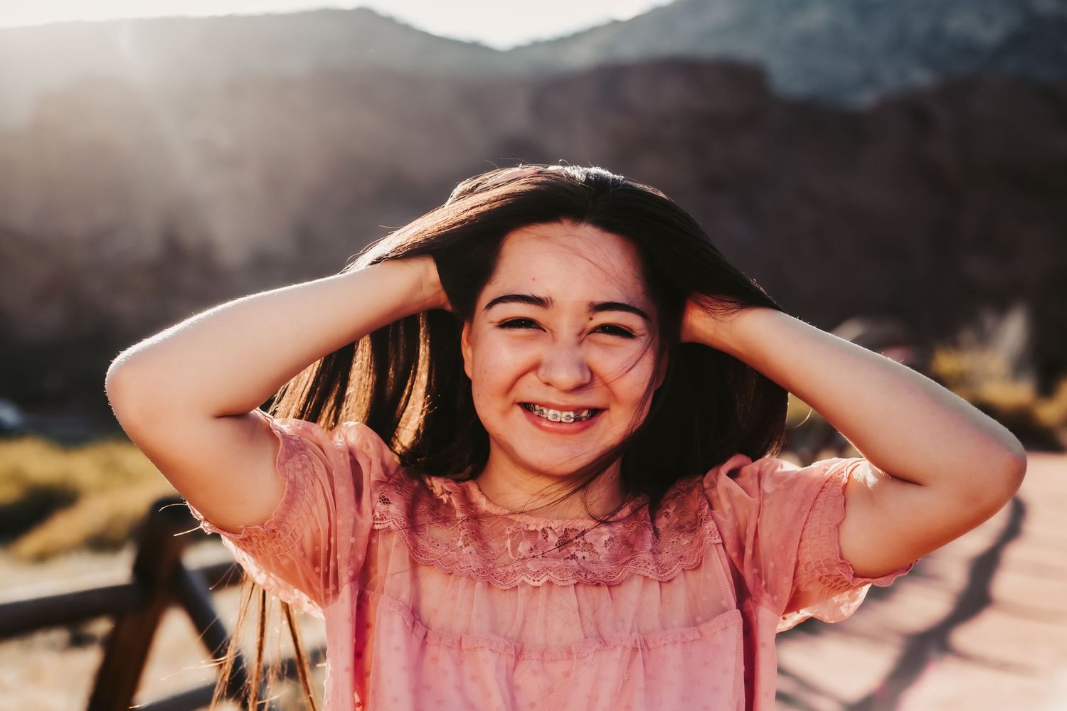 Person in pink shirt laughing joyfully with hands in hair against mountain backdrop at sunset.