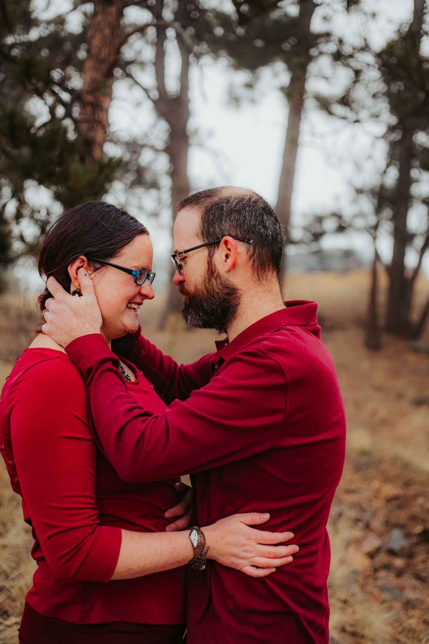 A couple wearing matching red sweaters embraces tenderly on a forest trail.