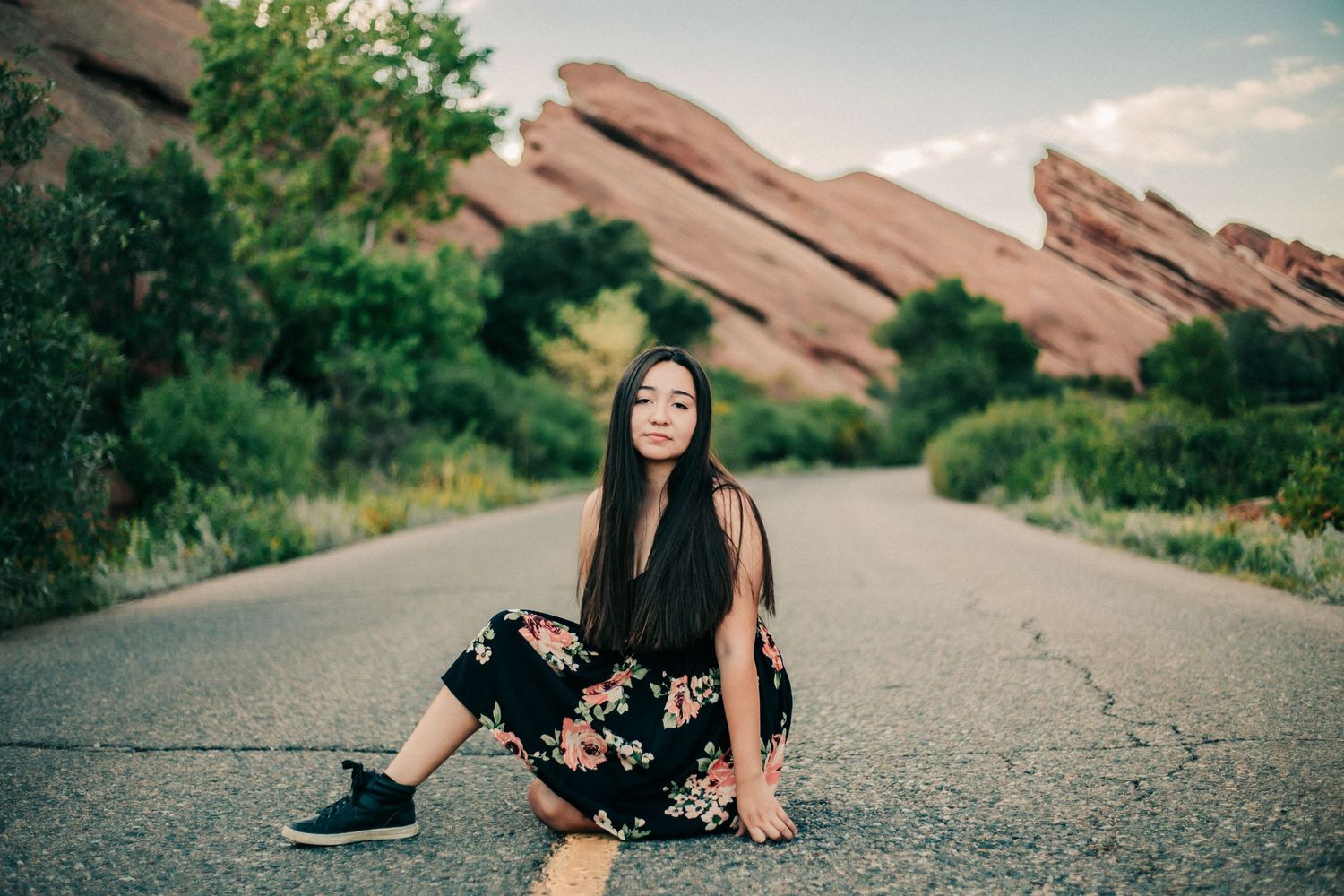 Woman in black floral dress sits on road with scenic red rock formations behind her.