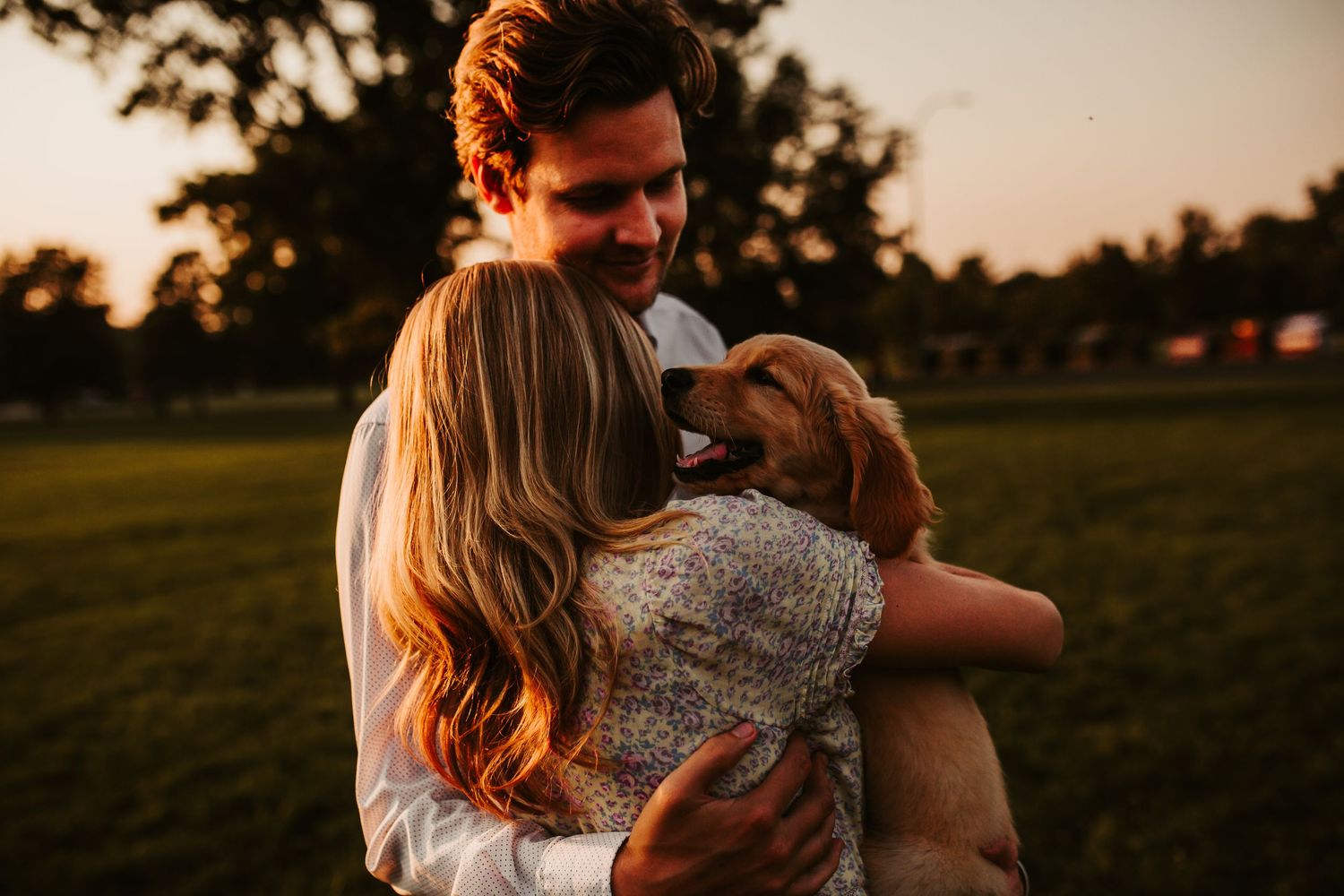 Couple embraces their golden retriever puppy during golden hour sunset in a grassy field.