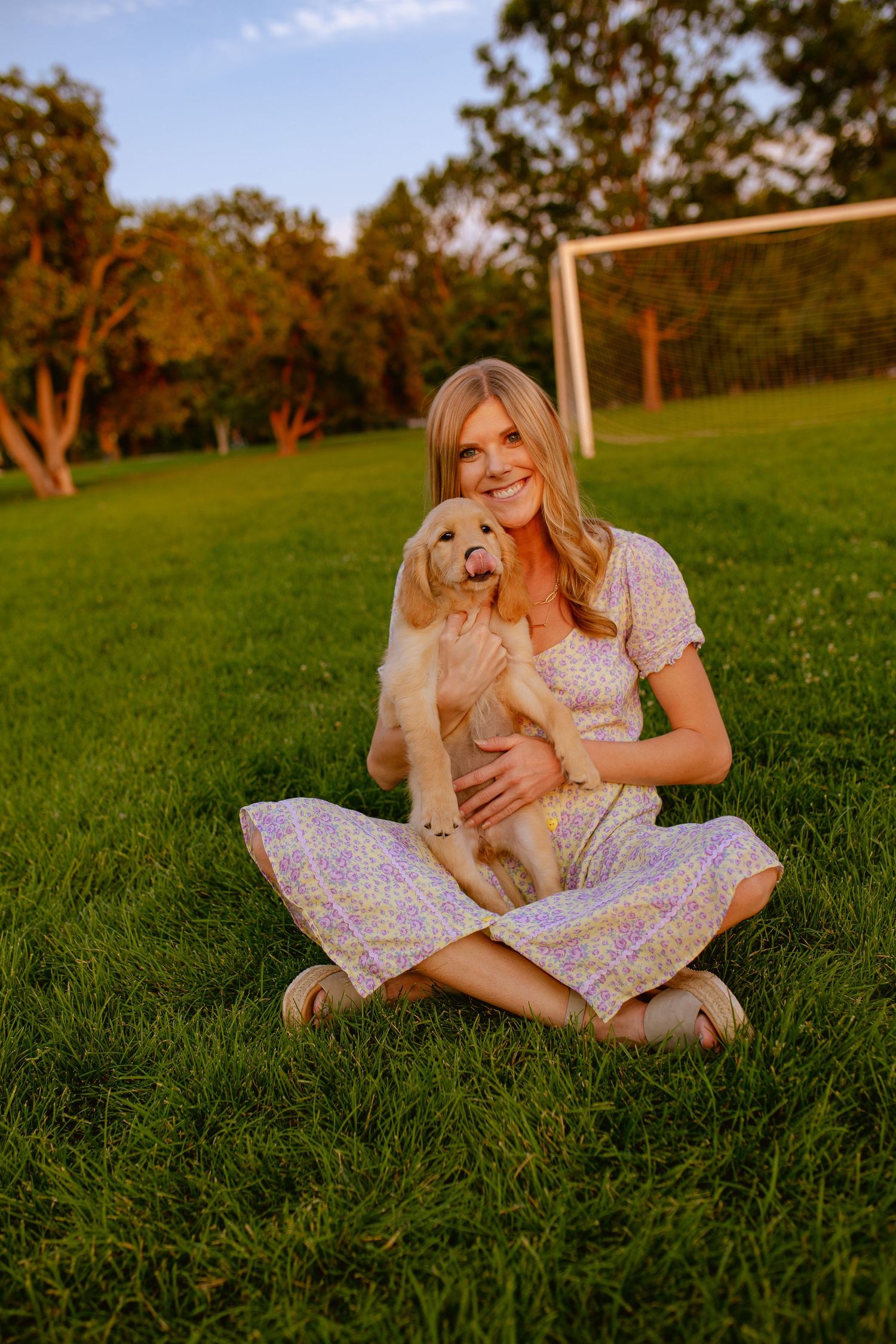 Woman sits cross-legged on grass with golden retriever puppy near soccer goal at sunset.