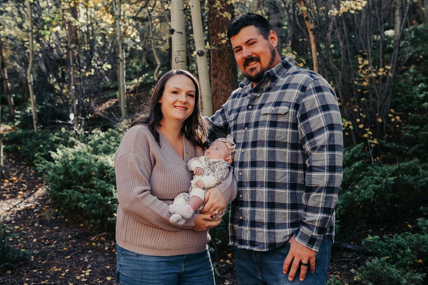 A couple holds their baby while standing in a wooded area with aspen trees.