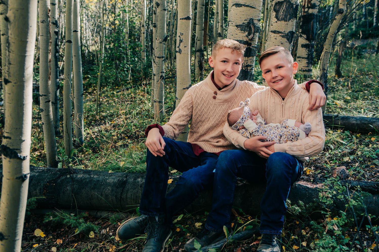 Two people sit on a log holding a baby in an aspen grove.