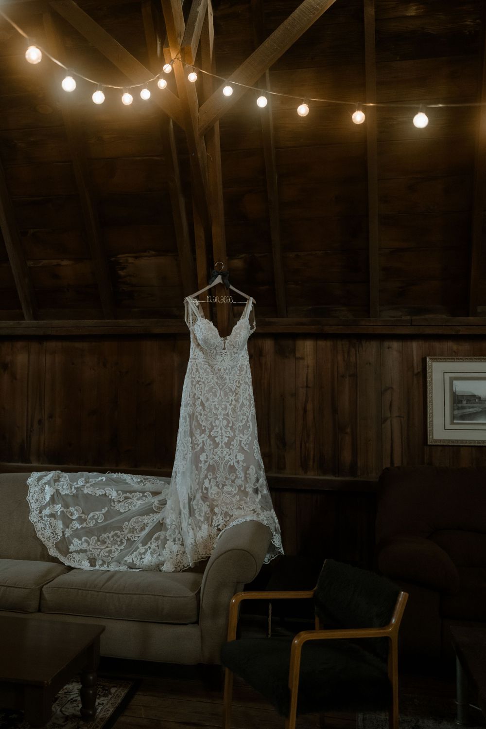 A delicate lace wedding dress hangs under string lights in a rustic wooden barn setting.