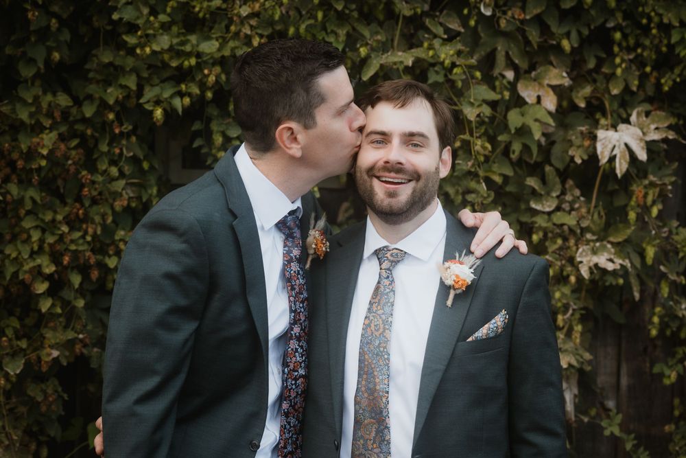 Two people in formal suits share a joyful moment against a leafy backdrop at a wedding celebration.