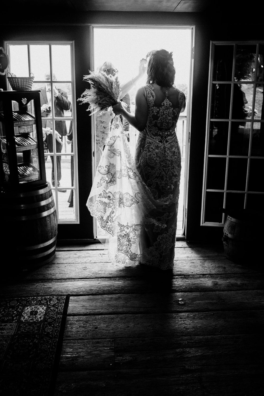 A dramatic silhouette of a bride in a lace dress standing by glass doors in black and white photography.