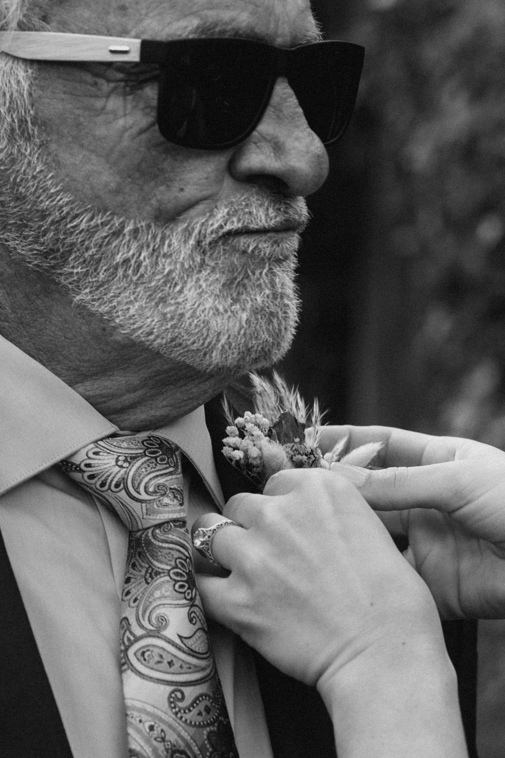 A close-up of a bearded man in sunglasses getting his patterned tie adjusted.