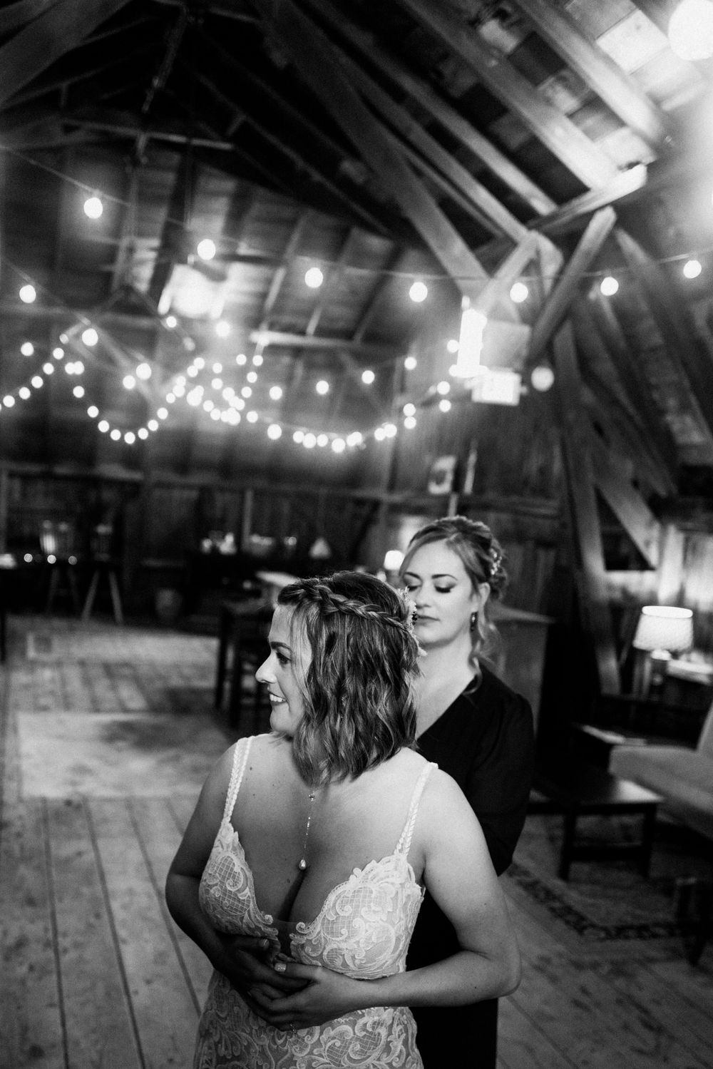 A black and white photo of a wedding dress fitting in a barn with twinkling lights overhead.