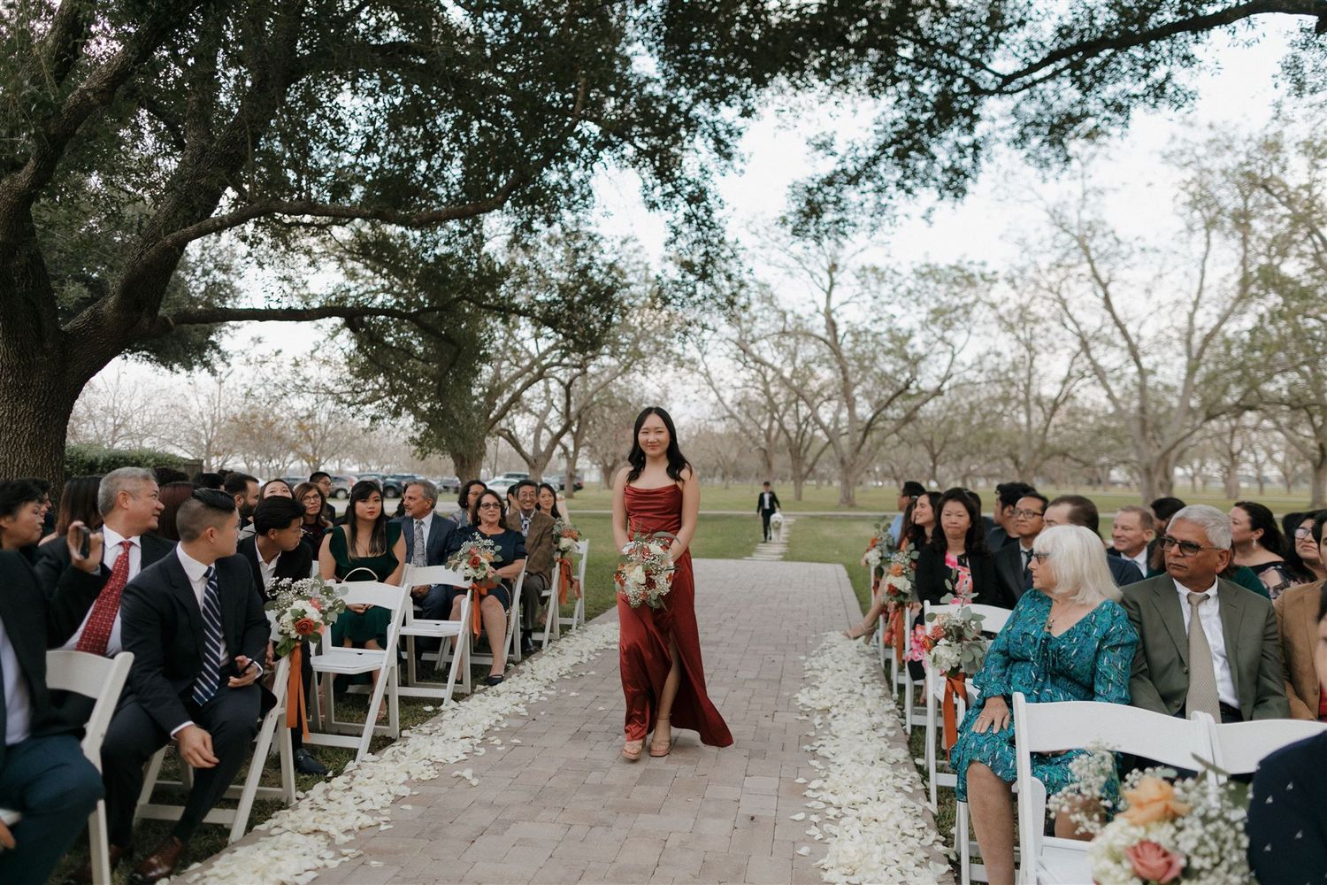 A figure in red walks down an outdoor aisle lined with white chairs and wedding guests.
