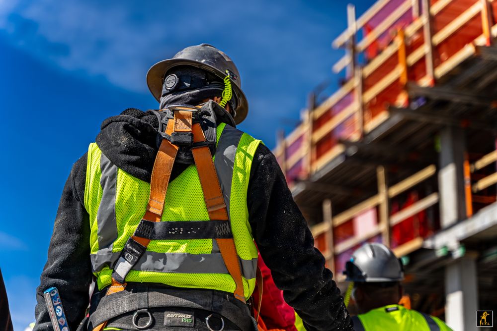 Construction worker wearing safety gear and harness on site during sunny day with building in background.