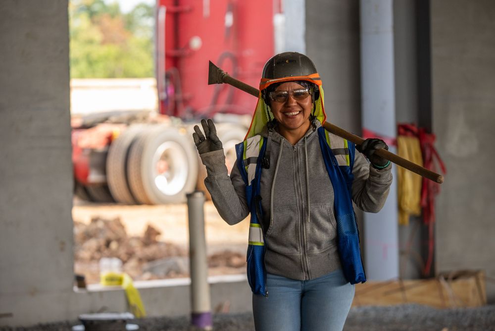 Construction worker carrying equipment while wearing protective gear at worksite.