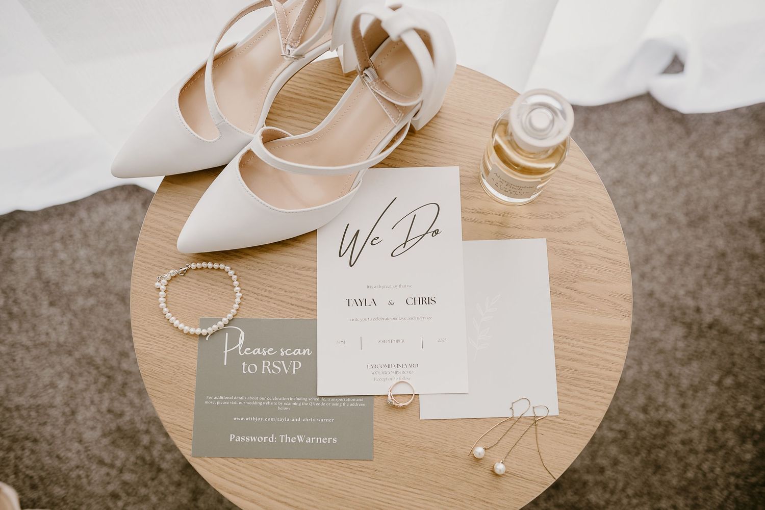 Wedding stationery and accessories including white heels and pearl jewelry on round wooden table.