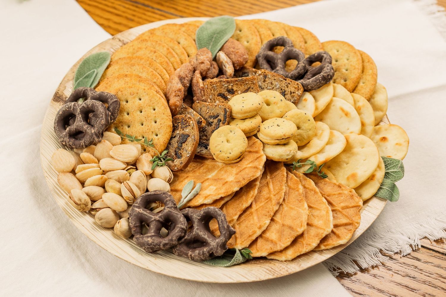An appetizer platter featuring round crackers, chocolate covered pretzels, and mixed nuts with decorative leaves.