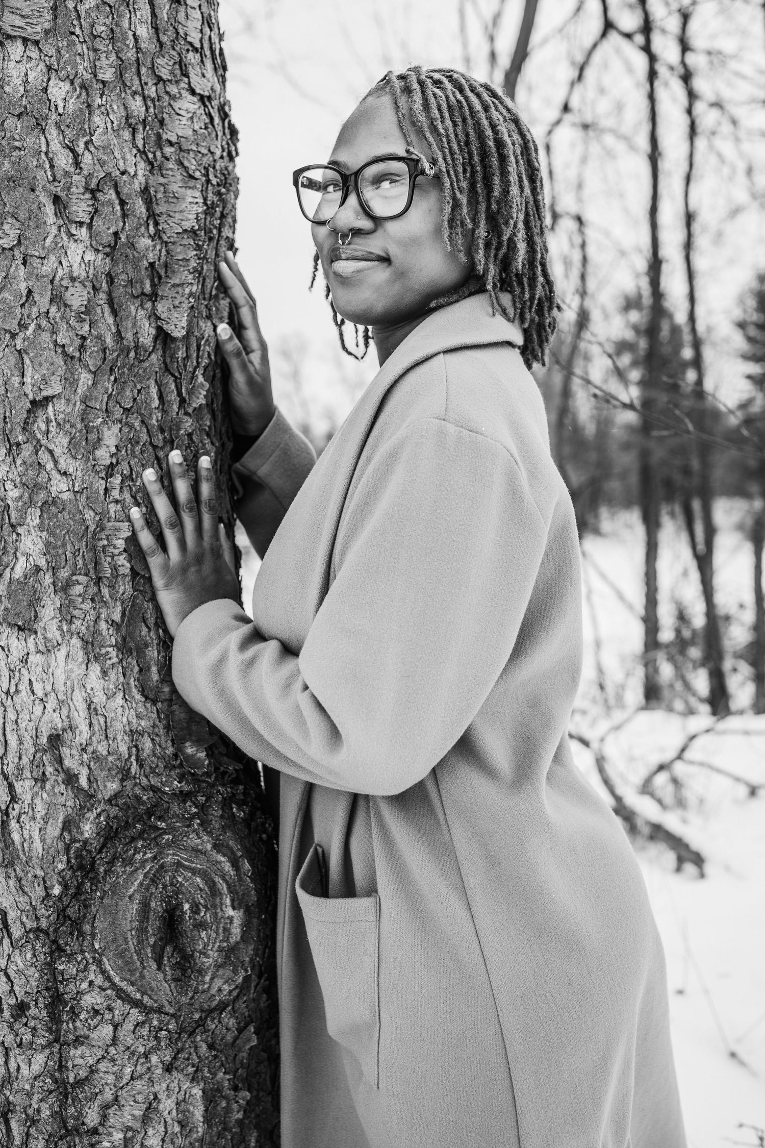 Black and white winter portrait leaning against tree trunk wearing beige coat.
