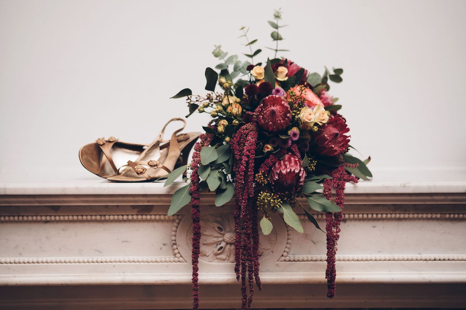 Wedding bouquet with burgundy and blush flowers rests on mantel beside golden bridal shoes.