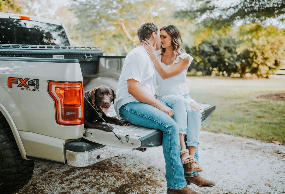 Couple sits on truck tailgate sharing a romantic moment with their chocolate lab dog outdoors.