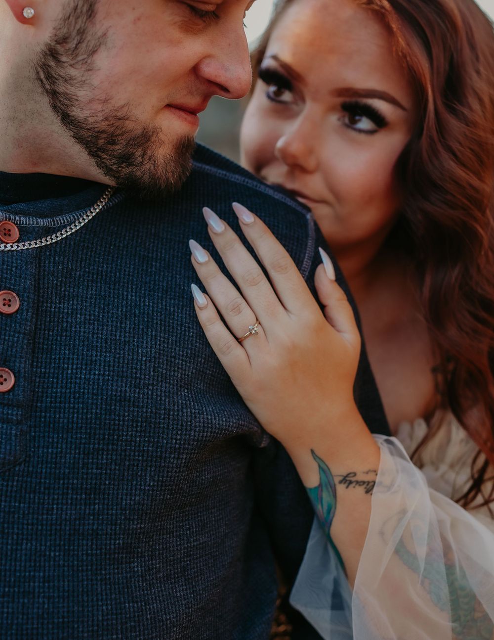 Couple in intimate moment, woman with red hair showing engagement ring and floral tattoo.