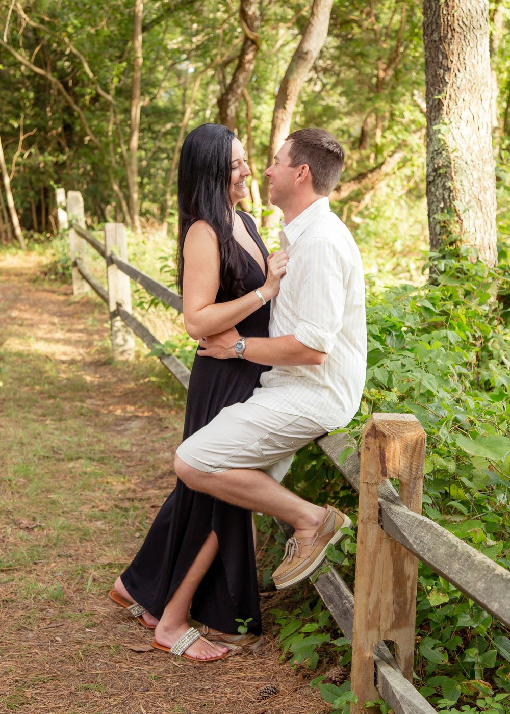 Couple embraces near a rustic wooden fence on a wooded trail, woman in black maxi dress.