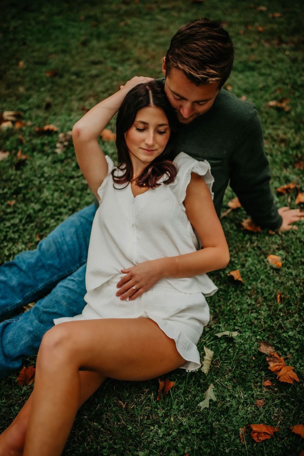 Couple sitting on grass in autumn park, woman in white dress leaning against man in green sweater.