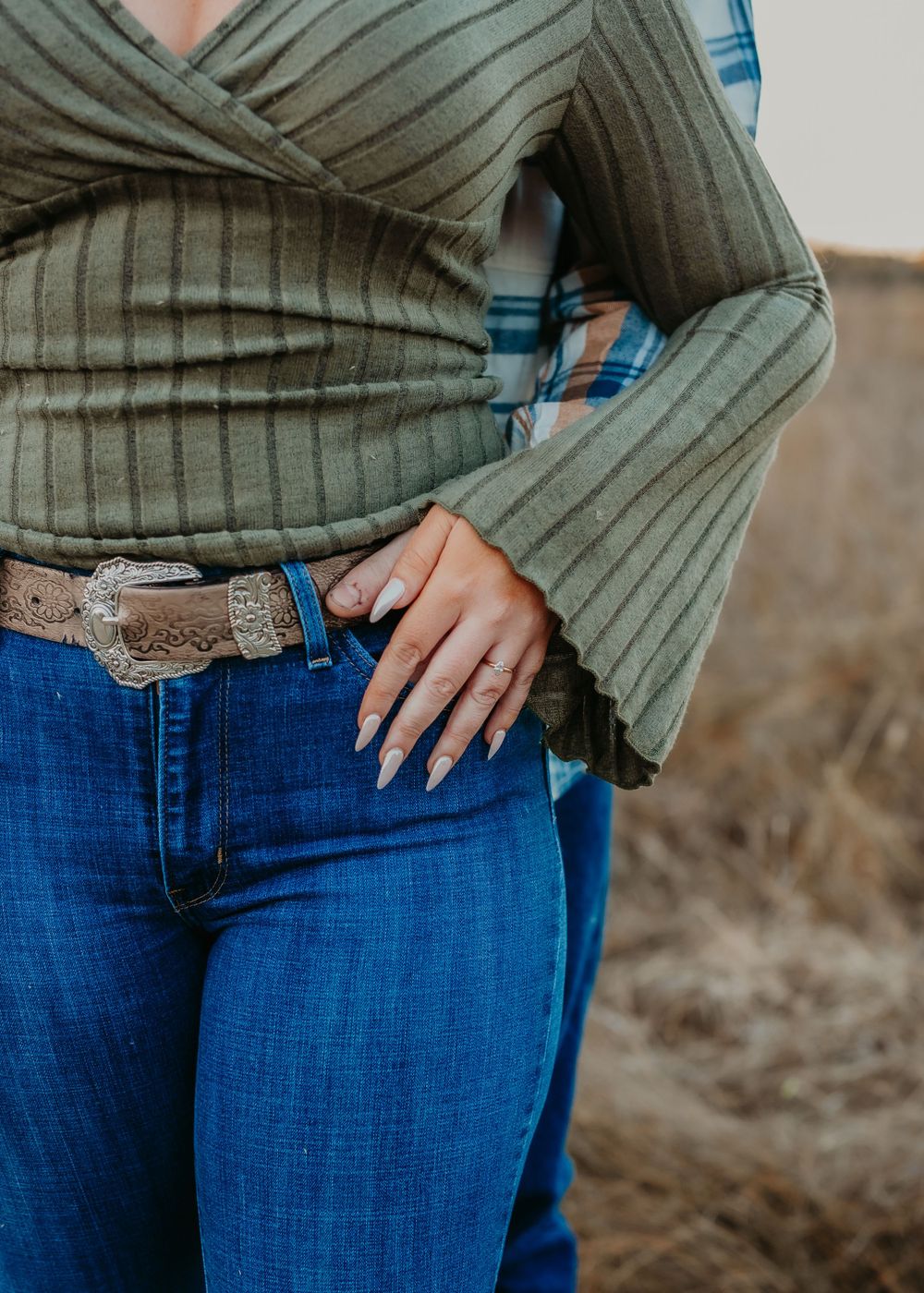 Close-up of woman in olive wrap top, western belt, and jeans showing engagement ring.