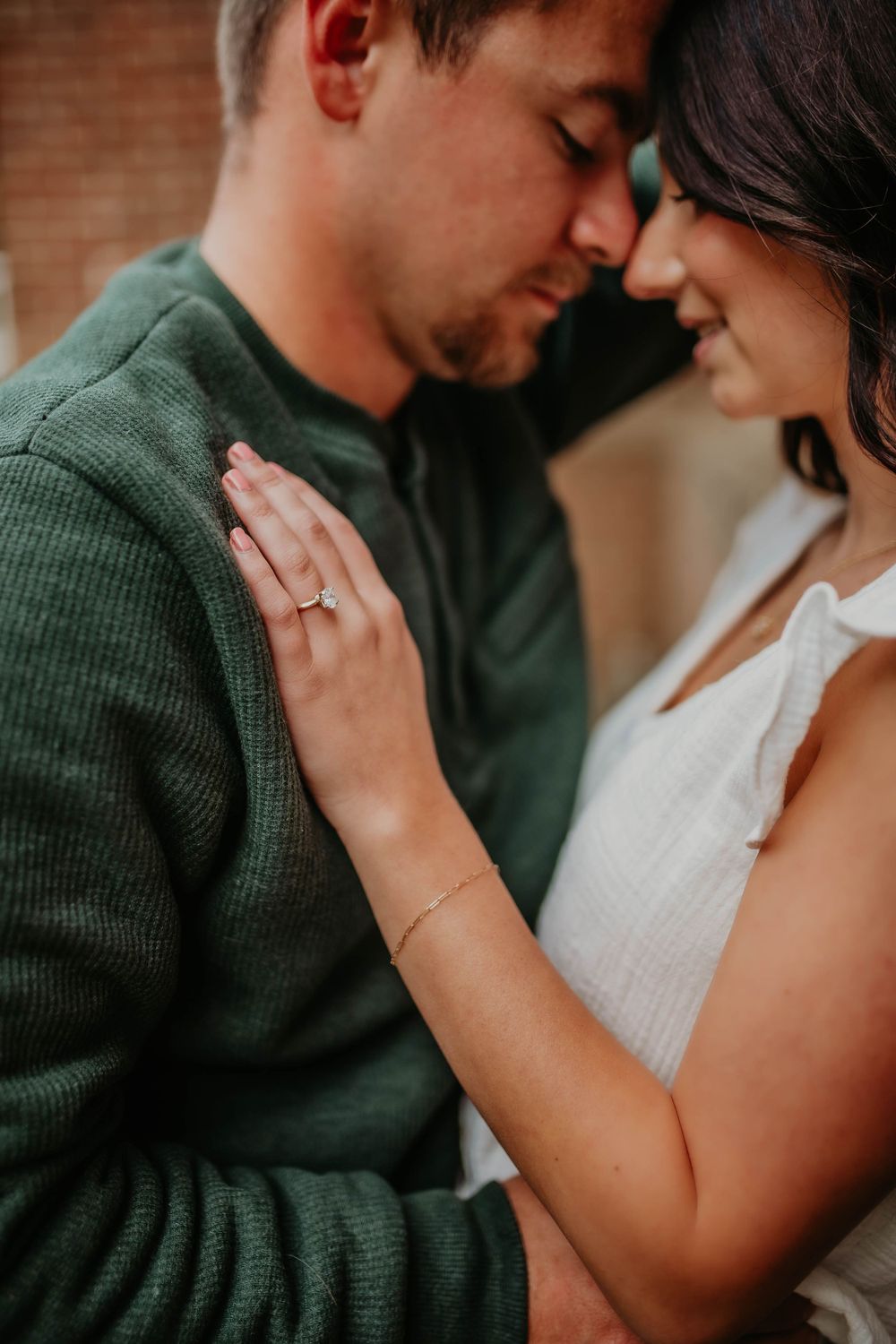 Intimate couple moment with woman in white dress showing engagement ring against green sweater.