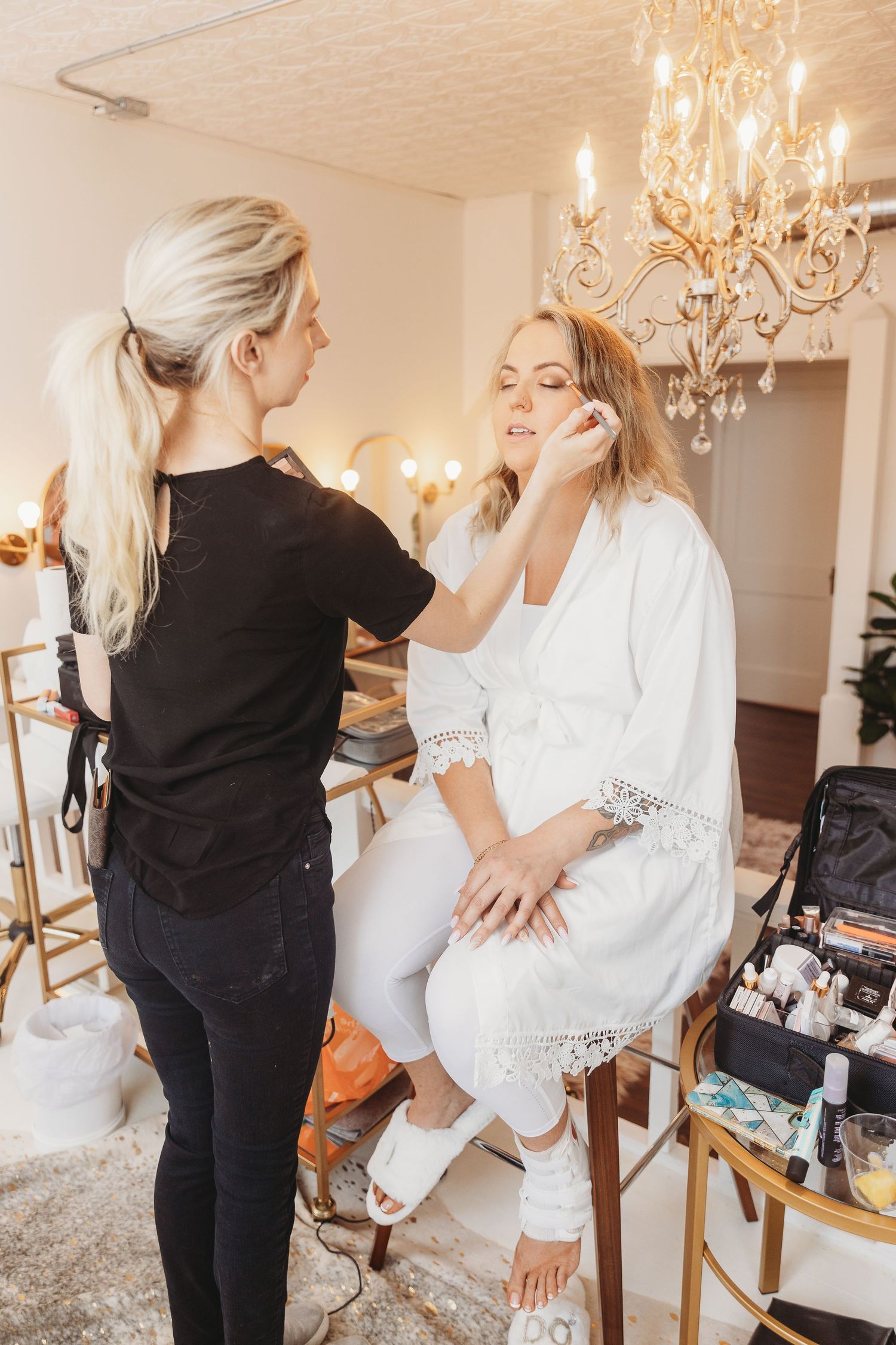A makeup artist applies cosmetics on a person wearing a white robe under an elegant crystal chandelier.
