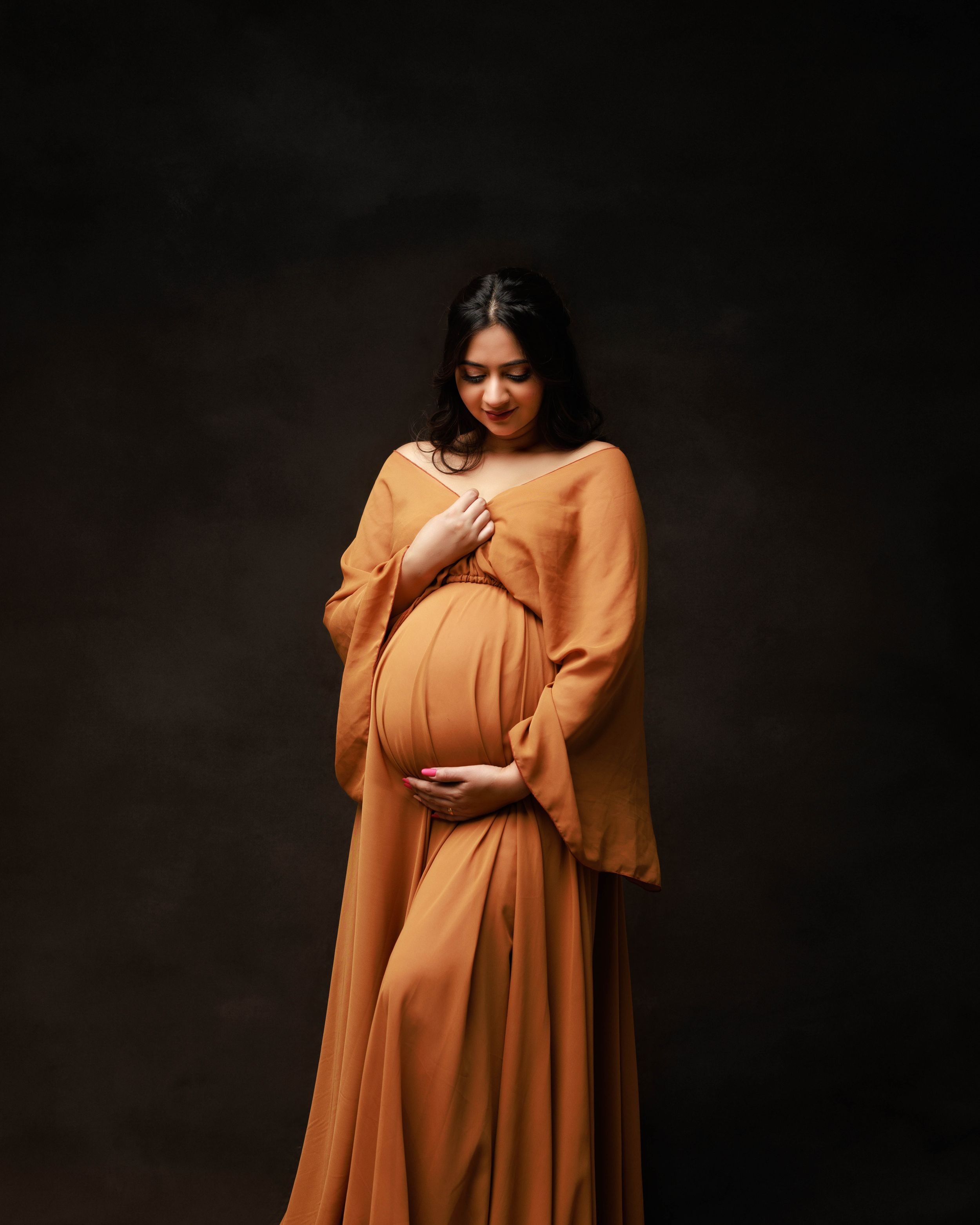 Expectant mother in a flowing orange dress poses against dark backdrop during maternity photoshoot.