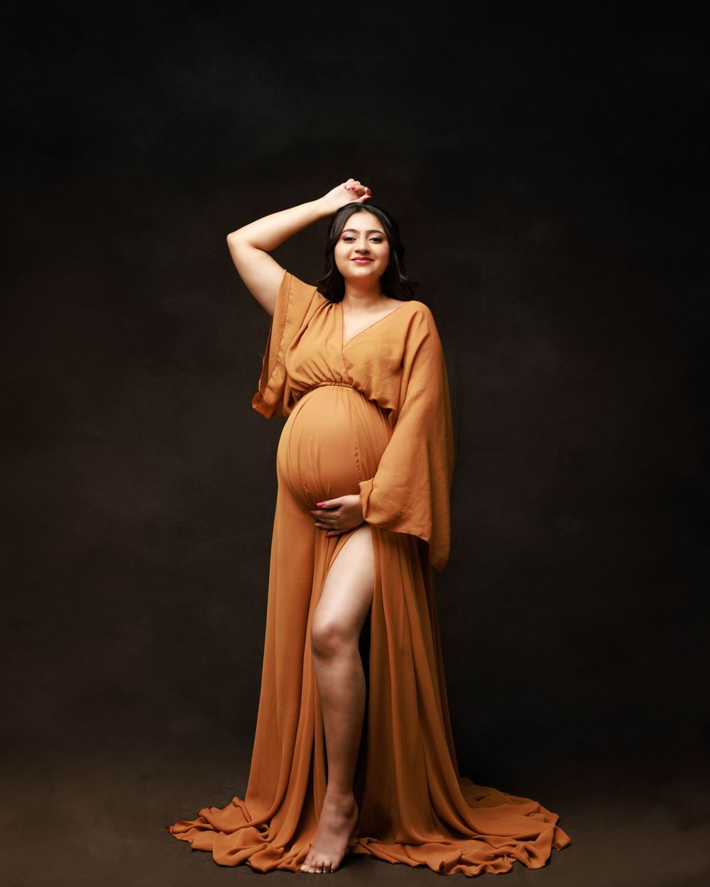 Elegant maternity portrait featuring a flowing orange gown with dramatic side slit against dark background.