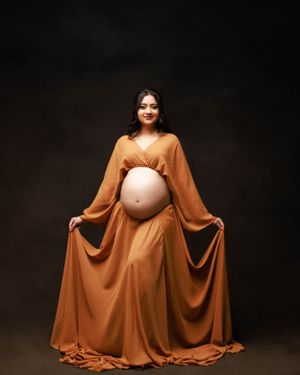 Dramatic maternity portrait with flowing orange dress and bell sleeves against dark background.