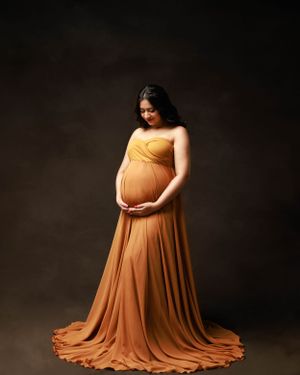 Maternity portrait with elegant orange floor-length gown against dark studio background.