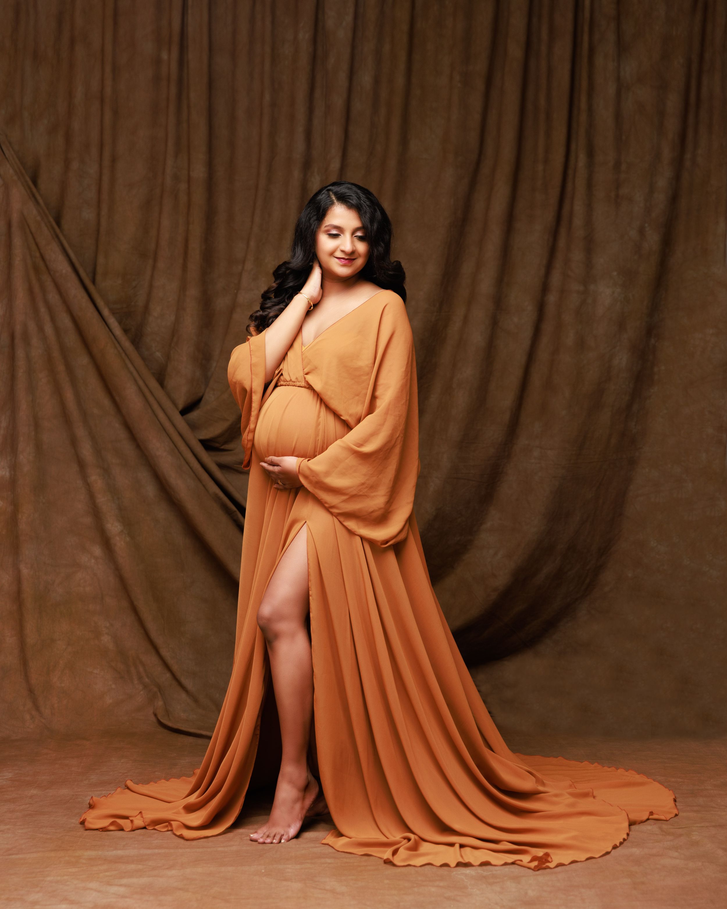 Elegant maternity pose in a floor-length orange gown with dramatic slit.