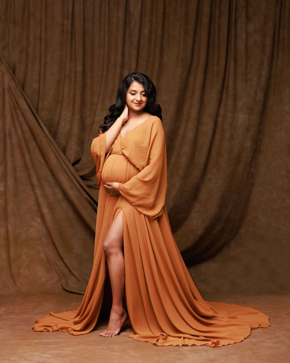 Elegant maternity pose in a floor-length orange gown with dramatic slit.