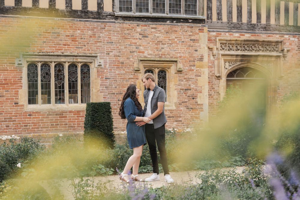 Couple embracing outside a historic brick building surrounded by blooming garden flowers.