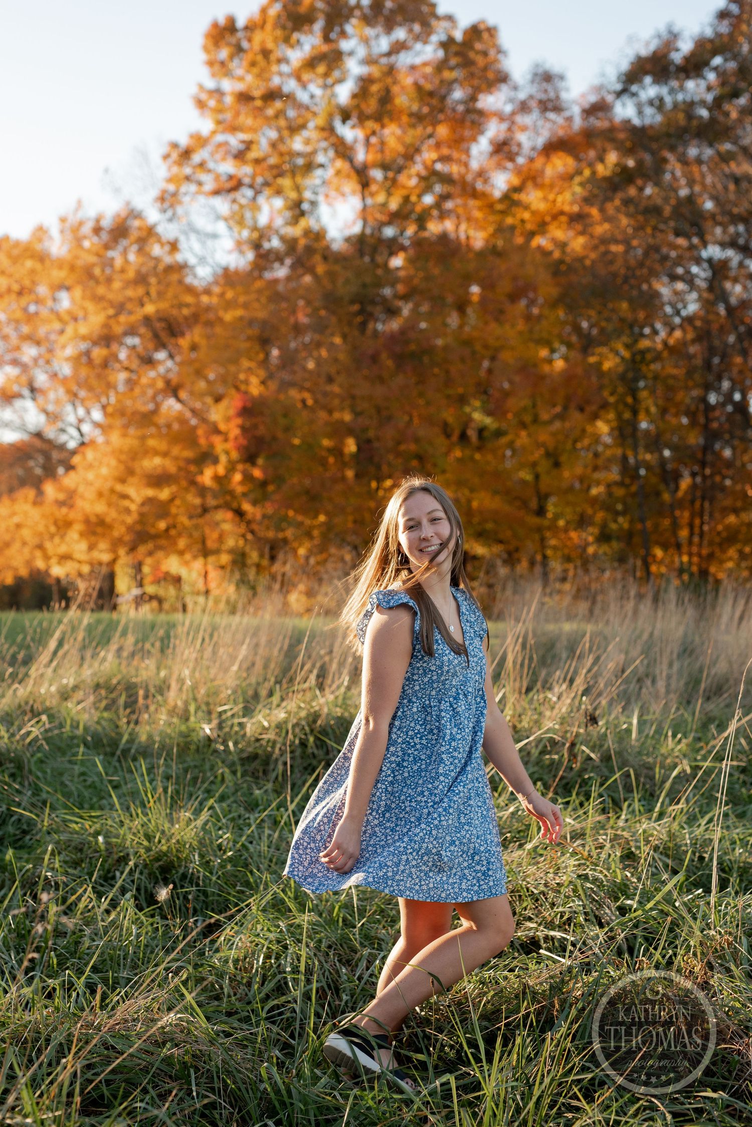 A flowy blue dress twirling in tall grass with autumn trees glowing in golden sunlight.