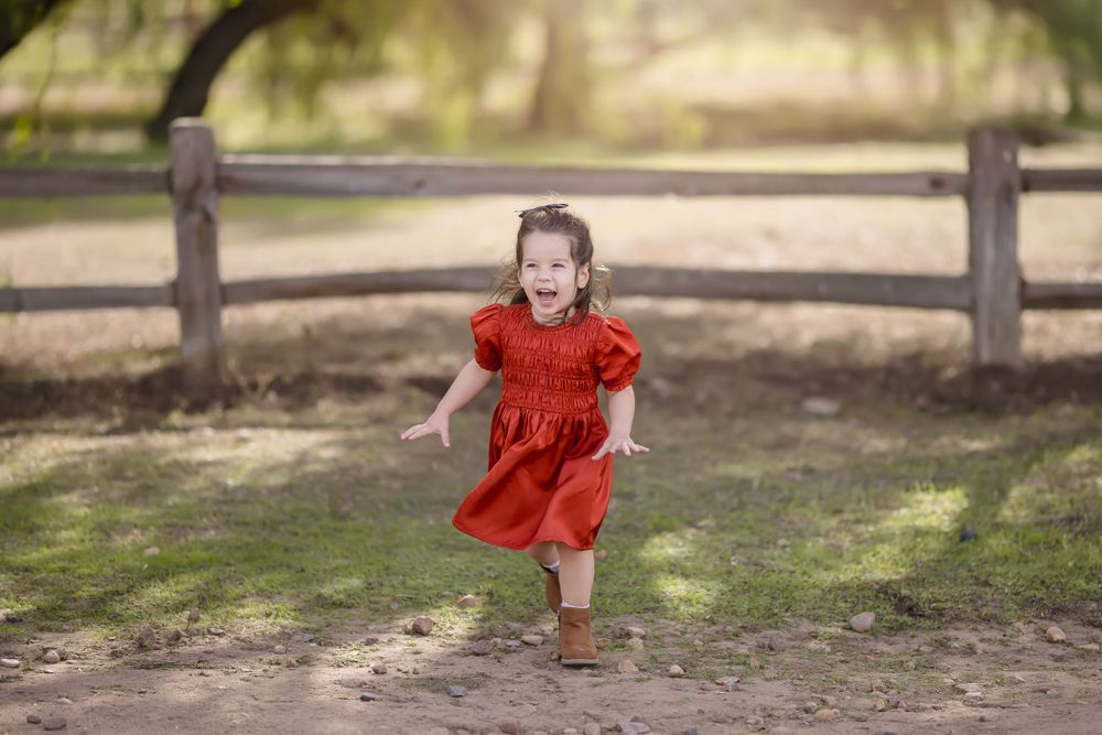 A small child in a red dress runs joyfully along a wooden fence in dappled sunlight.