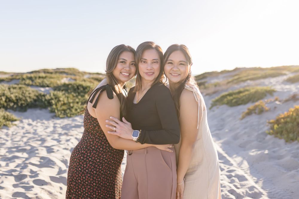 Three women embrace on a sunny beach with sand dunes in the background.