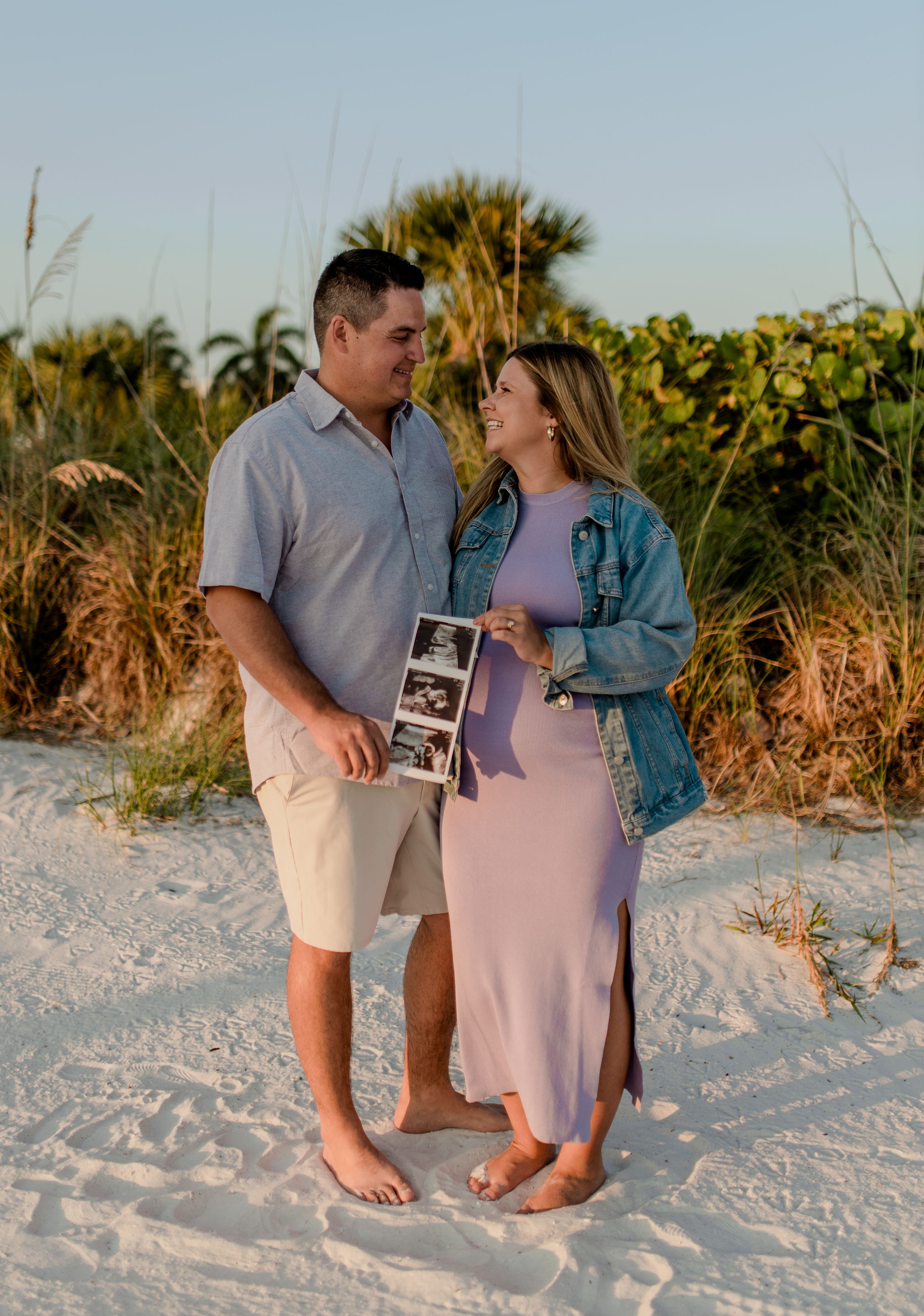 A couple standing in sunflowers holding an ultrasound photo during a sunset beach pregnancy announcement photoshoot.