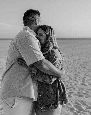 A couple embraces tenderly on a sandy beach in black and white.
