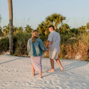 A couple walks barefoot together on a beach path surrounded by palm trees at sunset.