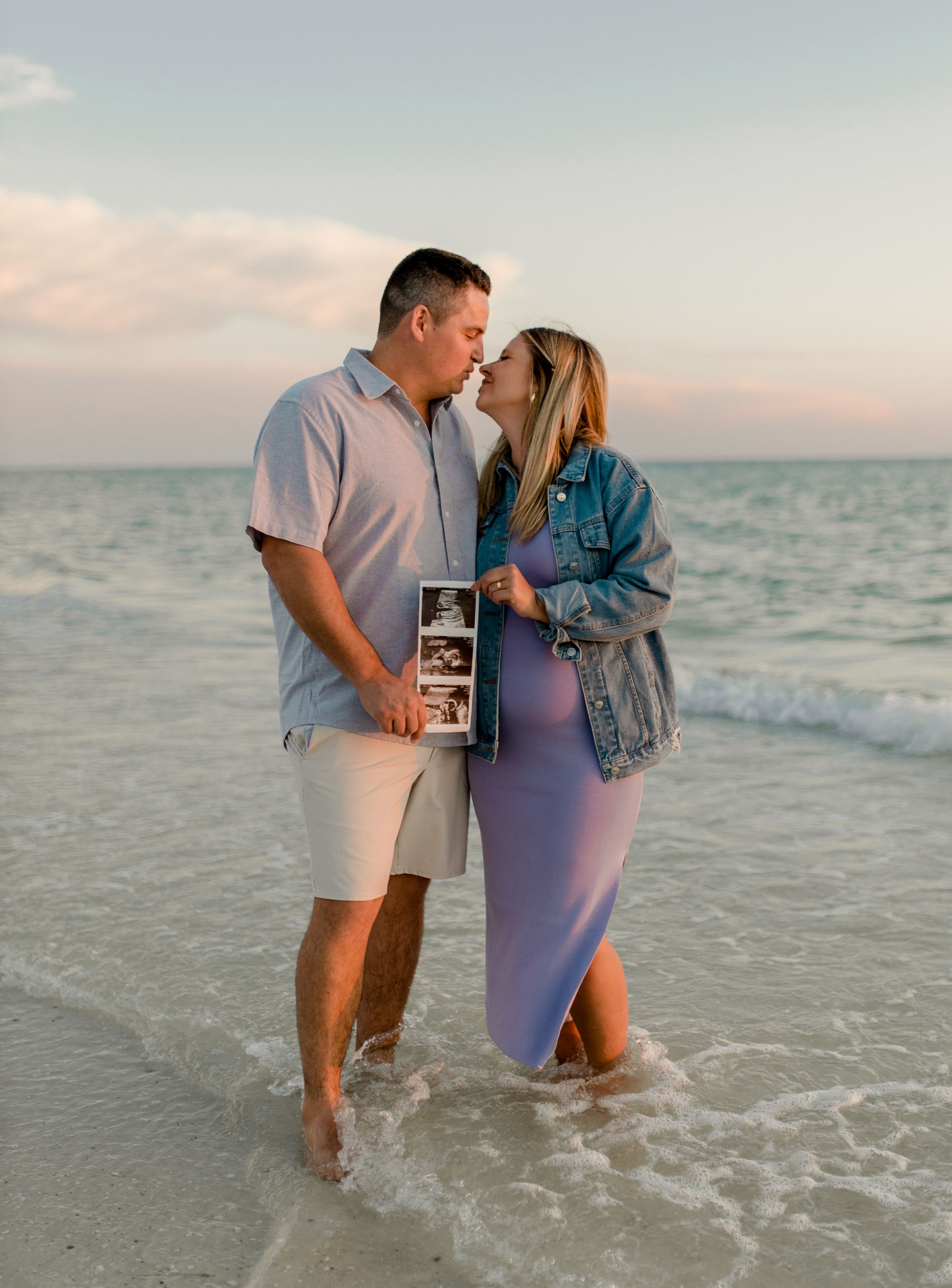 A couple sharing an intimate moment in the ocean waves while holding ultrasound photos.