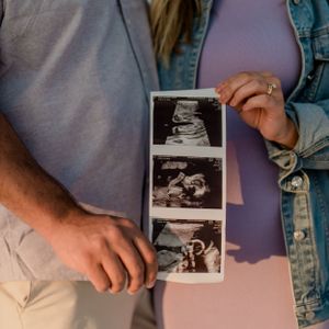 Hands hold ultrasound images against a pink dress and denim jacket background.